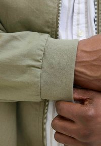 Khaki bomberjack met een mouw met ribgebreide manchetten, van katoen met een rits. Een hand houdt de manchet vast en onthult een wit gestreept shirt daaronder.