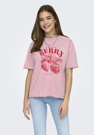 Jonge vrouw die glimlacht, draagt een oversized roze T-shirt met een rode aardbeienprint en lichtblauwe gescheurde jeans, poserend tegen een lichte achtergrond.