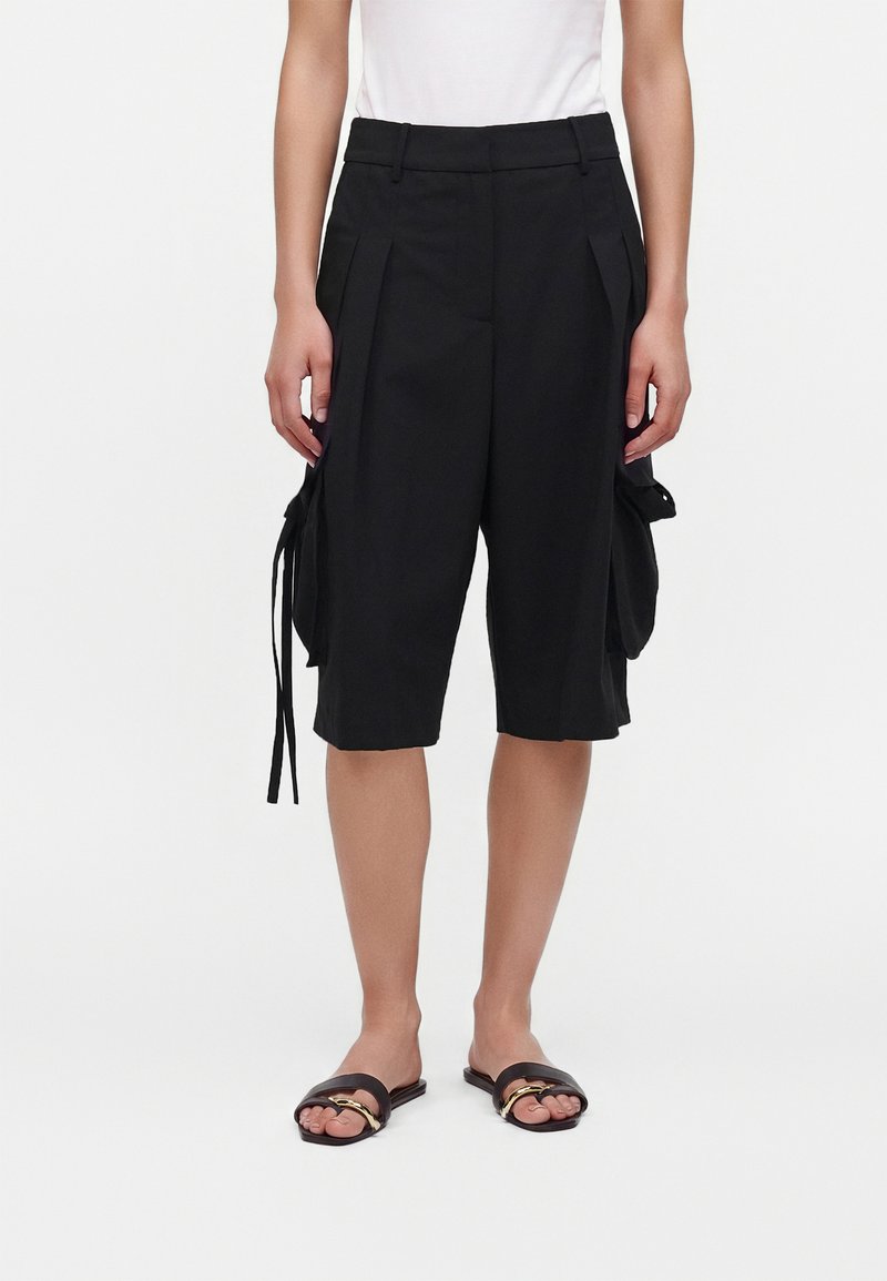 Personne portant un short cargo noir au genou avec des plis et des poches latérales, associé à des sandales slides noires et un haut blanc.