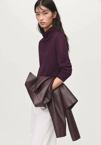 Suéter de cuello alto morado oscuro en un tejido suave, con mangas largas y un corte relajado, combinado con una chaqueta de cuero burdeos drapeada.