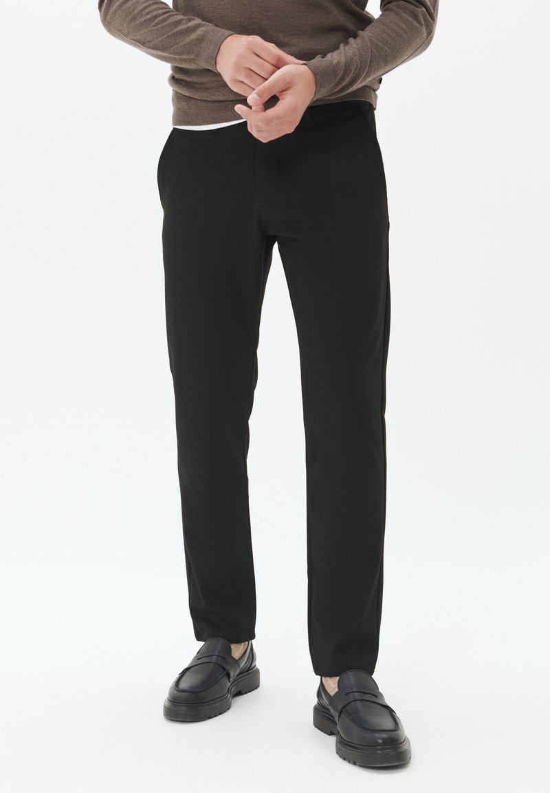 Pantalones negros ajustados con una textura suave, que presentan un corte clásico y bolsillos frontales, combinados con mocasines oscuros y un suéter en tonos neutros.