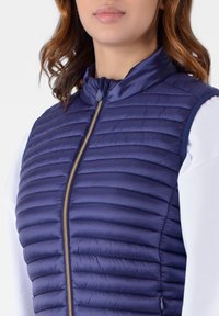 Gilet imbottito blu navy con motivo a righe orizzontali e colletto alto. Presenta una chiusura frontale con zip ed è indossato sopra una camicia bianca a maniche lunghe.