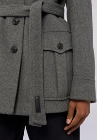 Šedý vlněný kabát ze směsového materiálu s dvouřadým zapínáním, zavazovacím pasem a přední kapsou s klopou. Texturovaná tkanina zvyšuje teplo.