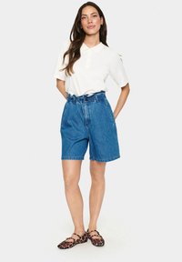 Jeansshorts mit hoher Taille, hellblauer Farbe und gerafftem Bund. Getragen mit einem weißen Poloshirt und flachen Schuhen im Leopardenmuster.
