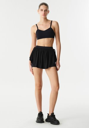 Reggiseno sportivo nero con spalline regolabili abbinato a una mini gonna nera a campana. La modella indossa scarpe da ginnastica nere. Texture del tessuto liscia.