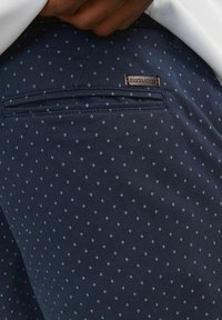 Pantalones marinos con un pequeño patrón geométrico blanco, con un bolsillo trasero y una etiqueta de marca visible en la cintura. Tejido texturizado.