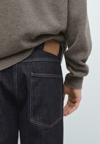 Dunkle Denim-Jeans mit strukturierter Oberfläche, ausgestattet mit Gesäßtaschen und einem braunen Lederetikett am Bund, kombiniert mit einem grauen Strickpullover.