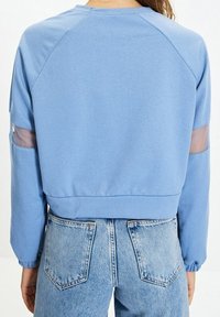 Světle modrý zkrácený svetr s raglánovými rukávy, který má průhledné síťované panely na rukávech a elastické manžety. Nosený s džíny s vysokým pasem.