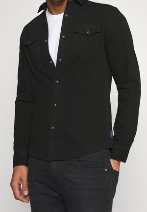 Homme portant une chemise noire à manches longues boutonnée sur un t-shirt blanc avec un jean noir, main dans la poche, sur un fond uni.