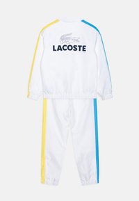 Tuta bianca con strisce gialle e blu. Presenta un alto colletto e polsini elastici. Logo "LACOSTE" e grafica di coccodrillo sul retro.