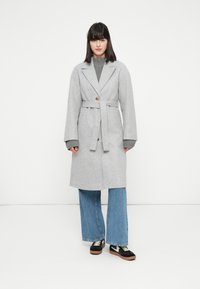 Donna in un cappotto grigio chiaro con cintura sopra un maglione grigio, abbinato a jeans blu a gamba larga e sneaker nere, in piedi su un pavimento bianco.