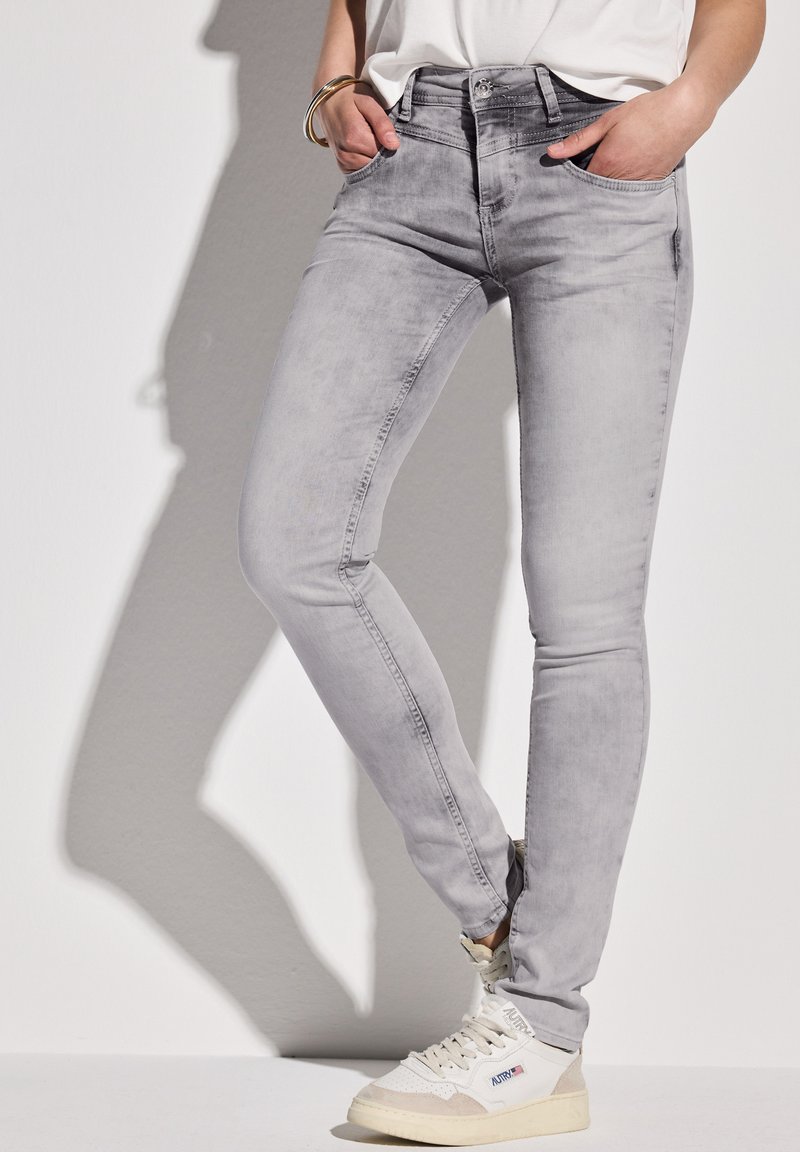 Vaqueros ajustados de denim gris con un acabado desgastado, que cuentan con bolsillos frontales y un cierre de botón, combinados con zapatillas altas blancas.