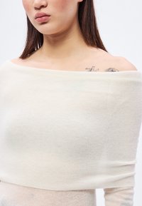 Krémový pletený top s odhalenými ramenami, jemná textúra, ľahká látka, priliehavý strih, jemný rebrovaný vzor a mierne priehľadný vzhľad.