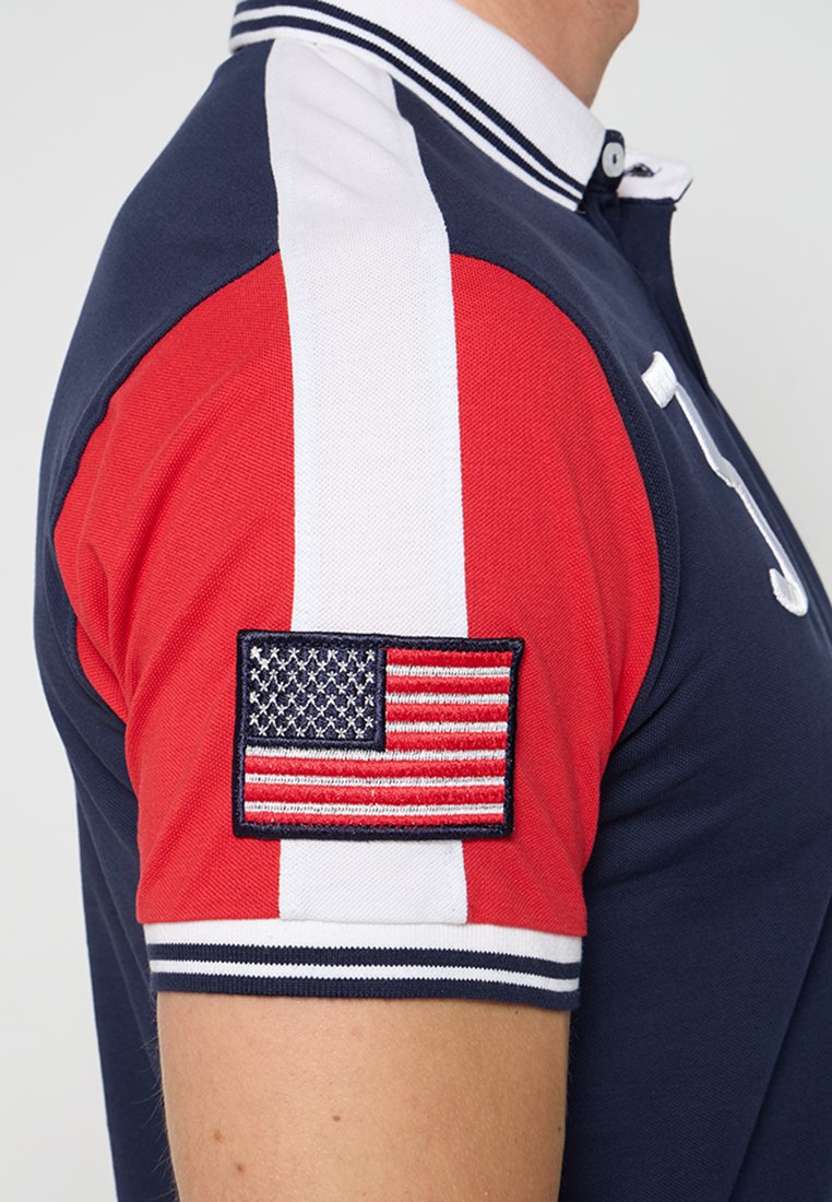 Un homme portant un polo bleu marine, rouge et blanc avec un patch brodé représentant le drapeau américain sur la manche rouge.