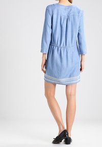 Femme portant une robe en denim bleu clair avec de la broderie blanche près de l'ourlet et des manches trois-quarts, associée à des chaussures plates à clous noires.