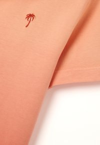 Camisa de manga corta en suave tela color durazno, con un pequeño diseño bordado de una palmera roja, con dobladillo limpio y textura suave.