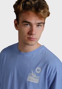 Ljusblå bomullst-shirt med rund hals, korta ärmar och en grafisk logo på bröstet med vågor och en elefantikon.