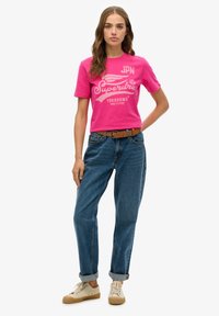 T-shirt en coton rose avec un impression graphique blanche, associé à un jean en denim bleu et des baskets beige. Le jean a des ourlets retroussés et une ceinture marron.