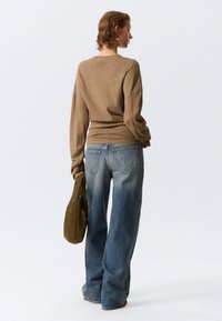 Καφέ μπλούζα με μακριά μανίκια, σε χοντρή γραμμή, συνδυασμένη με ανοιχτό μπλε τζιν παντελόνι με φαρδιά μπατζάκια. Το μοντέλο κρατάει μια πράσινη τσάντα και έχει κοντά, κυματιστά μαλλιά.