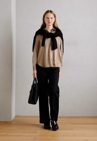 Beige button-up shirt, black trousers, oversized black sweater draped over shoulders, black handbag, chunky black shoes, neutral background.