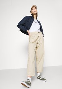 Veste bomber matelassée marine, T-shirt blanc avec logo, pantalon beige à cordon de serrage, baskets vertes et blanches, sur un fond neutre.