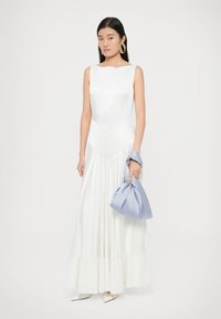 Balta be rankovių suknelė su plačia sijonu, turinti struktūruotą bodį ir subtilų blizgesį. Laiko šviesiai mėlyną audinio rankinę; paprastos auksinės auskarai.