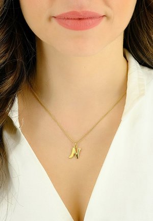 Collier en pendentif en or avec une initiale "N", accroché à une chaîne délicate, porté avec une chemise blanche à col.