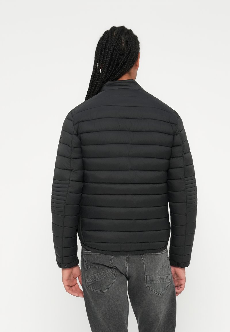 Doudoune noire avec un design matelassé, accents côtelés sur les manches et un col haut. Portée avec un jean gris, vue de dos.