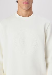 Sudadera color crema con un logo texturizado en relieve en el centro y acentos de laurel. Fabricada con una mezcla de algodón.