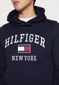 Sudadera con capucha de color azul marino que presenta "HILFIGER" y "NEW YORK" en letras blancas, con un logotipo rectangular en rojo, blanco y azul.