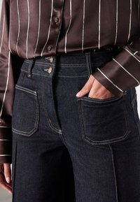 Jean en denim foncé à taille haute, avec deux poches avant, des coutures contrastantes, et un chemisier lisse à rayures en satin.