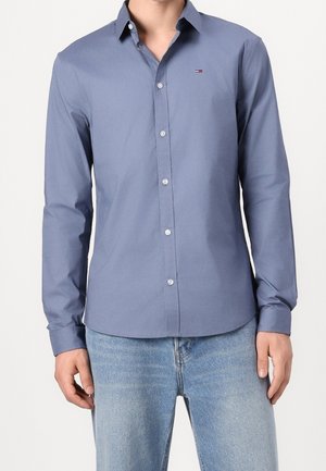 Mann trägt ein eng anliegendes, blaues Hemd mit langen Ärmeln und kleinem Logo auf der Brust sowie hellblaue Jeans.