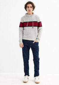 Sudadera de punto gris con una franja horizontal roja y negra, con cuello de cordón, combinada con jeans azul oscuro y zapatillas blancas.