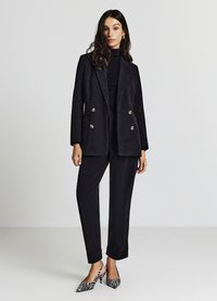 Blazer in corduroy nero con bottoni doppi, abbinato a pantaloni slim coordinati e décolleté a punta in bianco e nero a righe.