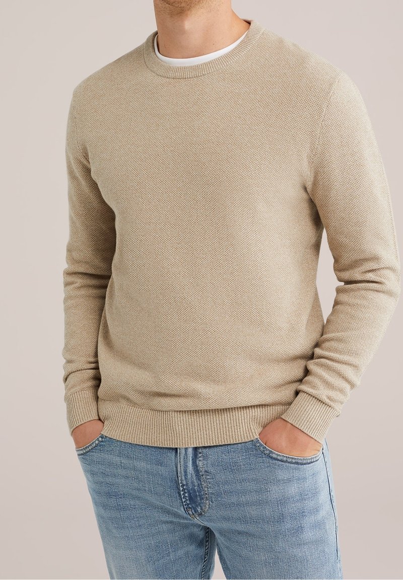 Homme portant un pull beige texturé à col rond et un jean bleu clair, mains dans les poches, sur un fond neutre.