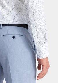 Camicia bianca con piccoli pois blu, polsini bottonati, abbinata a pantaloni azzurri chiari con tasca posteriore e un particolare con bottoni.