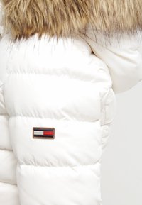 Vit puffjacka med huva av fuskpäls. Har vadderad textur och en liten, rektangulär logotyp i marinblått, röd och vitt.