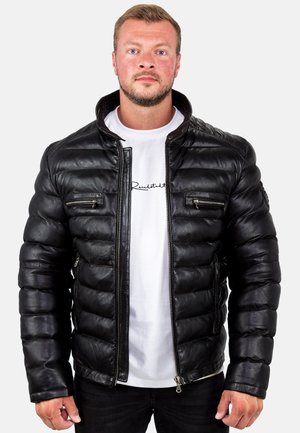 Man wearing a black padded leather jacket over a white t-shirt with black text, standing against a plain light background.