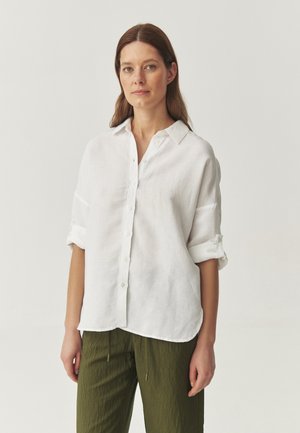 Camisa blanca de lino con frente abotonado, mangas enrollables y dobladillo curvado, combinada con pantalones de textura verde oliva.