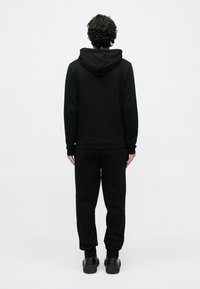 Muž s kučeravými vlasmi stojí čelom dozadu, má na sebe čiernu mikinu s kapucňou, čierne tepláky a čierne tenisky na jednoduchom svetlom pozadí.