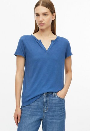 Frau mit glattem hellbraunem Haar, die ein blaues T-Shirt mit kurzen Ärmeln und V-Ausschnitt sowie blaue Jeans trägt, mit einer Hand in der Tasche.