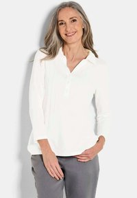 Weiße Bluse mit langen Ärmeln, Knopfleiste und Kragen, die vertikale Falten und eine glatte, weiche Textur aufweist. Getragen mit grauen Hosen.