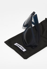 Námornícke modré slnečné okuliare s matným povrchom a tmavými šošovkami, odstavené na čiernom mäkkom puzdre s bielym štítkom "ARNETTE".