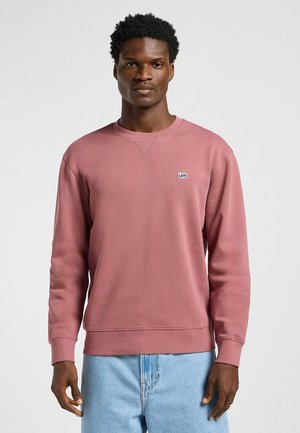 Różowy bawełniany sweatshirt z długimi rękawami, żebrowanym dekoltem i mankietami. Posiada małe niebieskie logo "Lee" na piersi. Połączony z jasnoniebieskimi dżinsami.