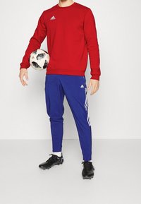 Rode sweatshirt, blauwe sportbroek met drie witte strepen, zwarte voetbalschoenen, met een zwart-wit gestructureerde voetbal.