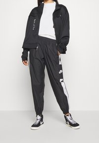 Black athletic jacket and pants with white side stripes. Jacket features zippers and branding; pants have side pockets and logo. Black sneakers.