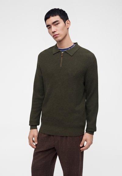 Jeune homme portant un pull zippé texturé vert foncé et un pantalon marron à carreaux, debout devant un fond clair uni.