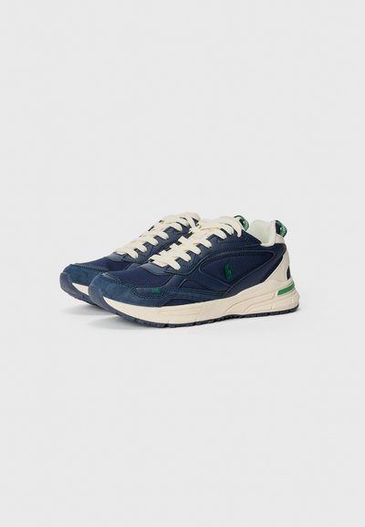 Tênis azul-marinho com parte superior em creme, apresentando um design texturizado, detalhes em verde e uma sola branca robusta. Fecho com atacadores.