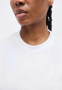 Κοντινό πλάνο του κάτω μέρους του προσώπου και του λαιμού ενός ατόμου που φοράει λευκό μπλουζάκι με στρογγυλή λαιμόκοψη και το λογότυπο "BORG" ανάγλυφο στο στήθος.