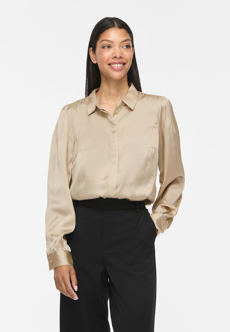 Femme souriante aux longs cheveux foncés portant un chemisier en satin beige à manches bouffantes et un pantalon noir taille haute, debout devant un fond blanc.